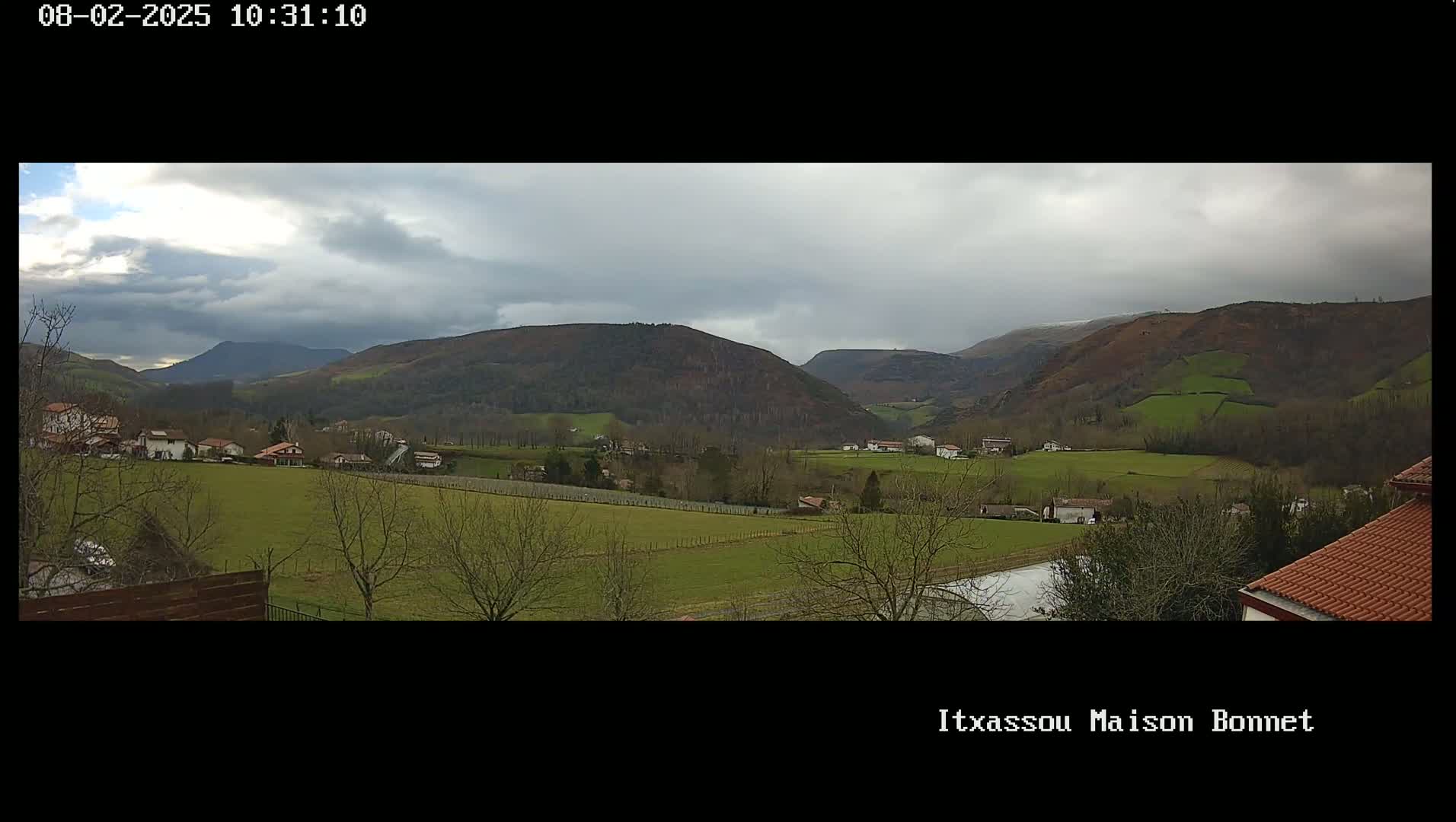 A panoramic view of a rural valley under a cloudy sky shows rolling green hills, sparsely scattered houses, and leafless trees.