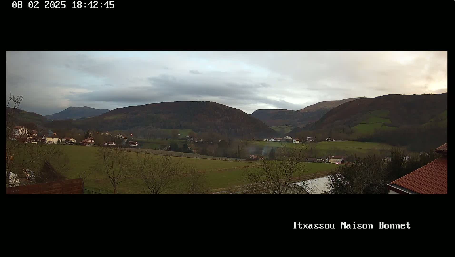 A panoramic view of a valley under a cloudy sky shows a small village nestled among green fields and dark, rolling hills.