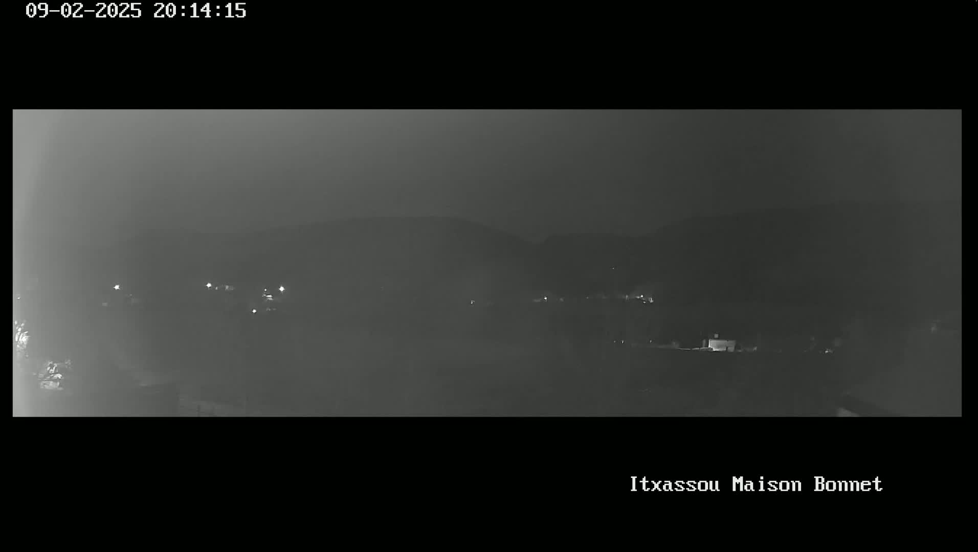 A nighttime, hazy view of a dark landscape with a few distant lights and hills in the background.