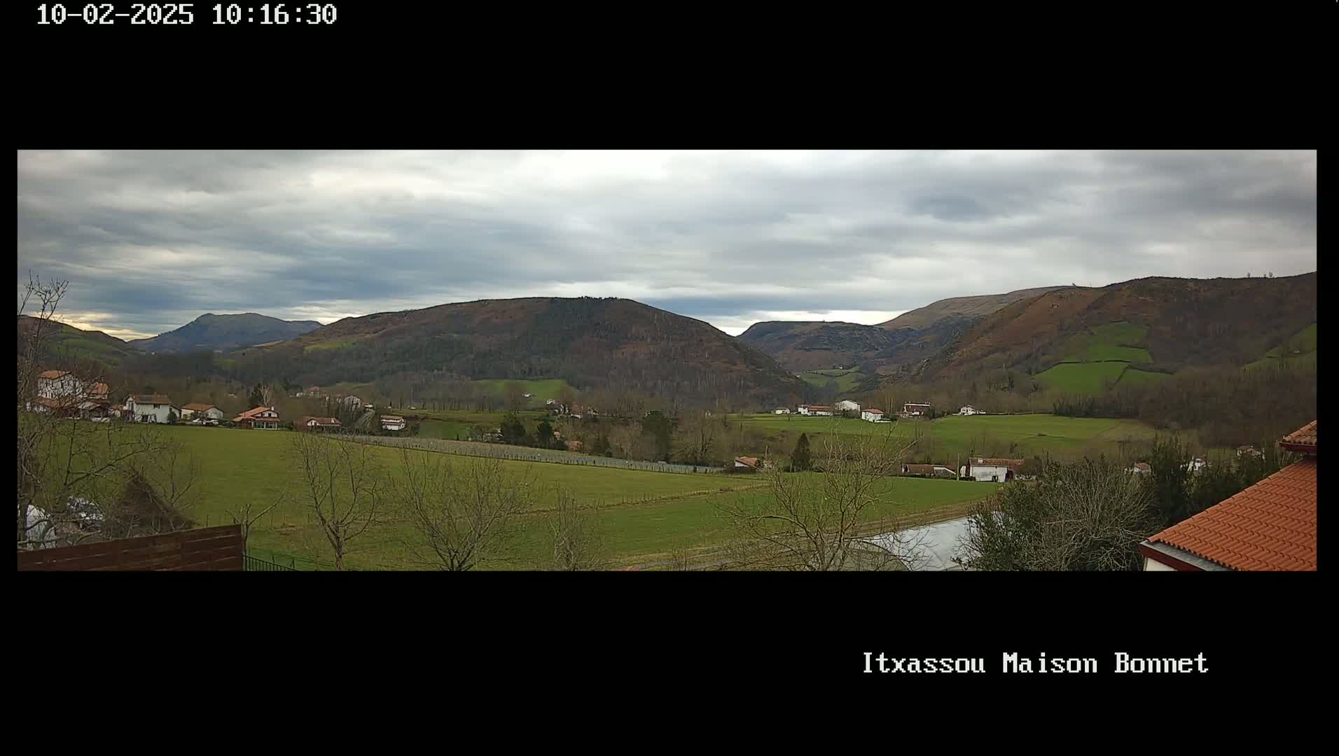 A panoramic view of a rural valley under a cloudy sky shows a scattering of houses and farmlands nestled among rolling green hills.