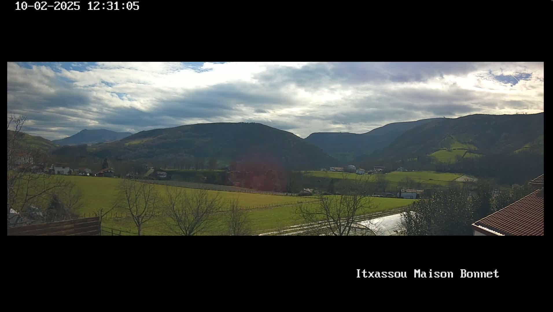 A partly cloudy day reveals a panoramic view of rolling green hills and farmland, with distant mountains under a mostly cloudy sky.