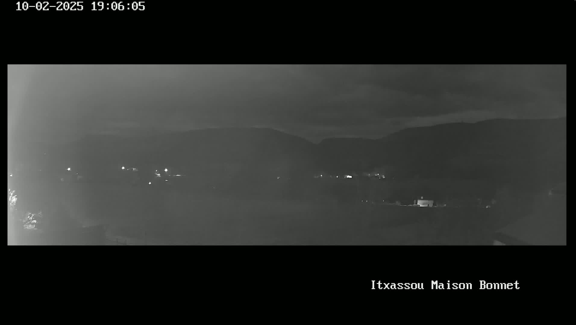 A nighttime, low-light grayscale image shows a dimly lit landscape with distant hills and a few scattered lights, possibly a small town, under a cloudy sky.