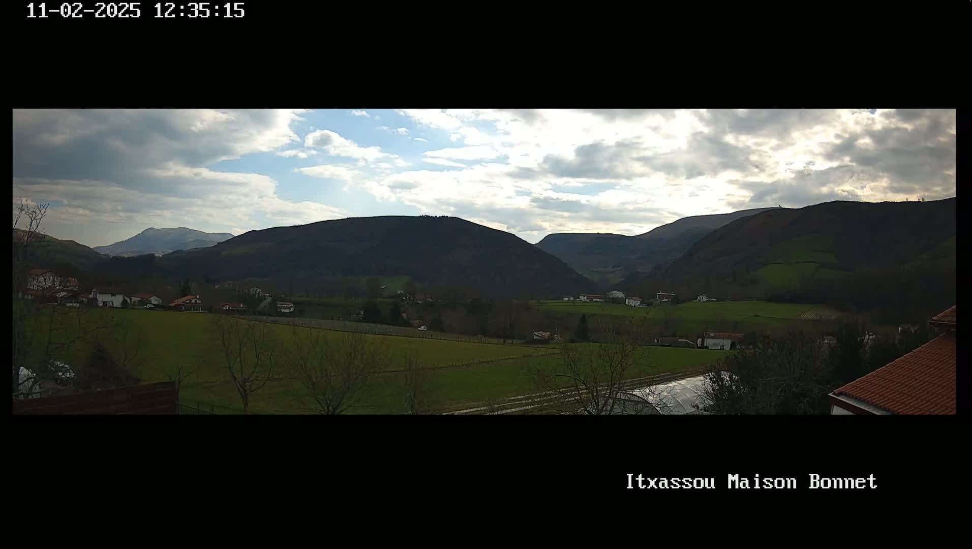 A partly cloudy day reveals a sprawling valley dotted with houses and farmlands, nestled among rolling green hills and mountains.