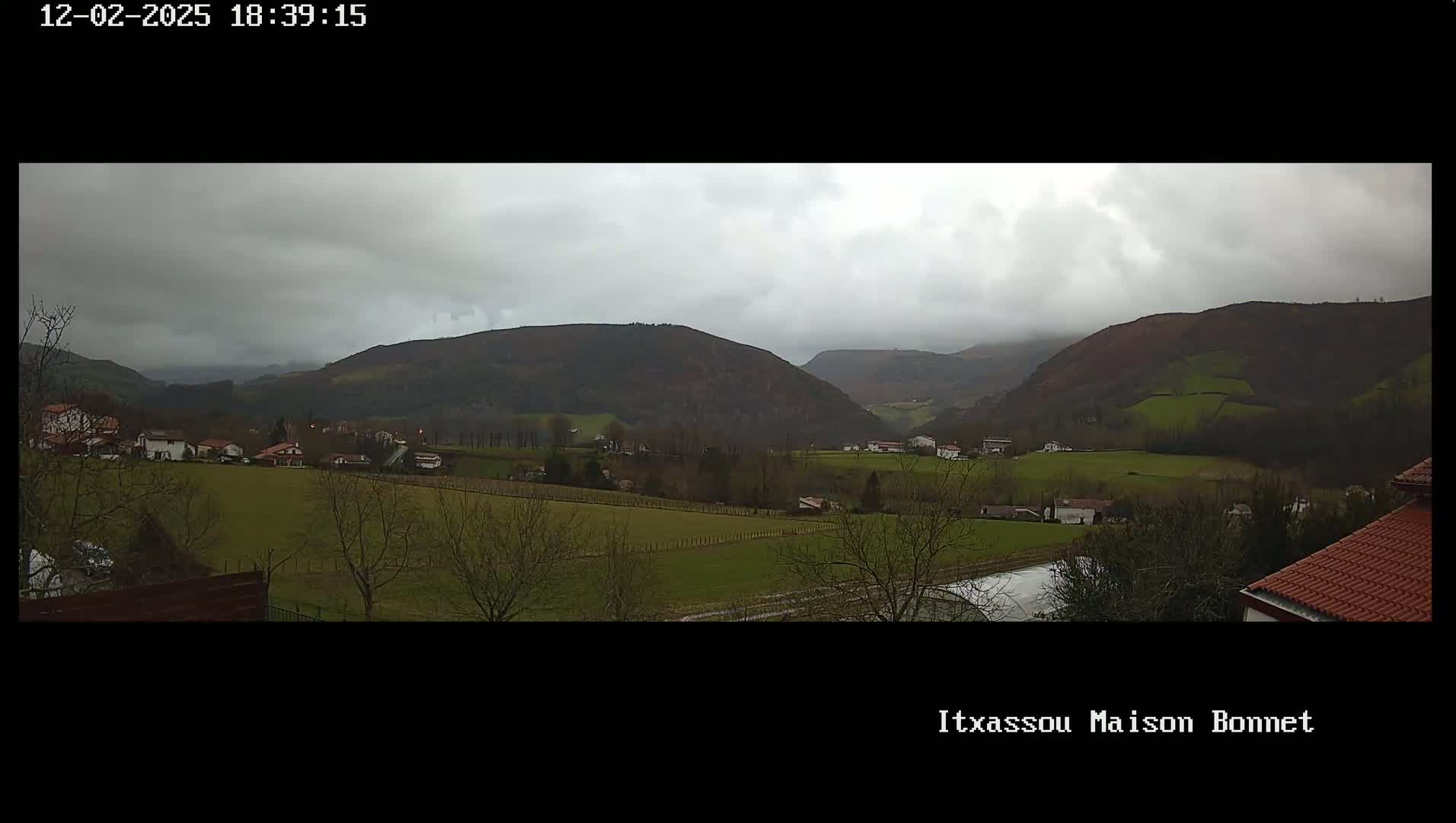 A panoramic view of a valley with overcast skies shows a small village nestled amongst green fields and rolling hills.