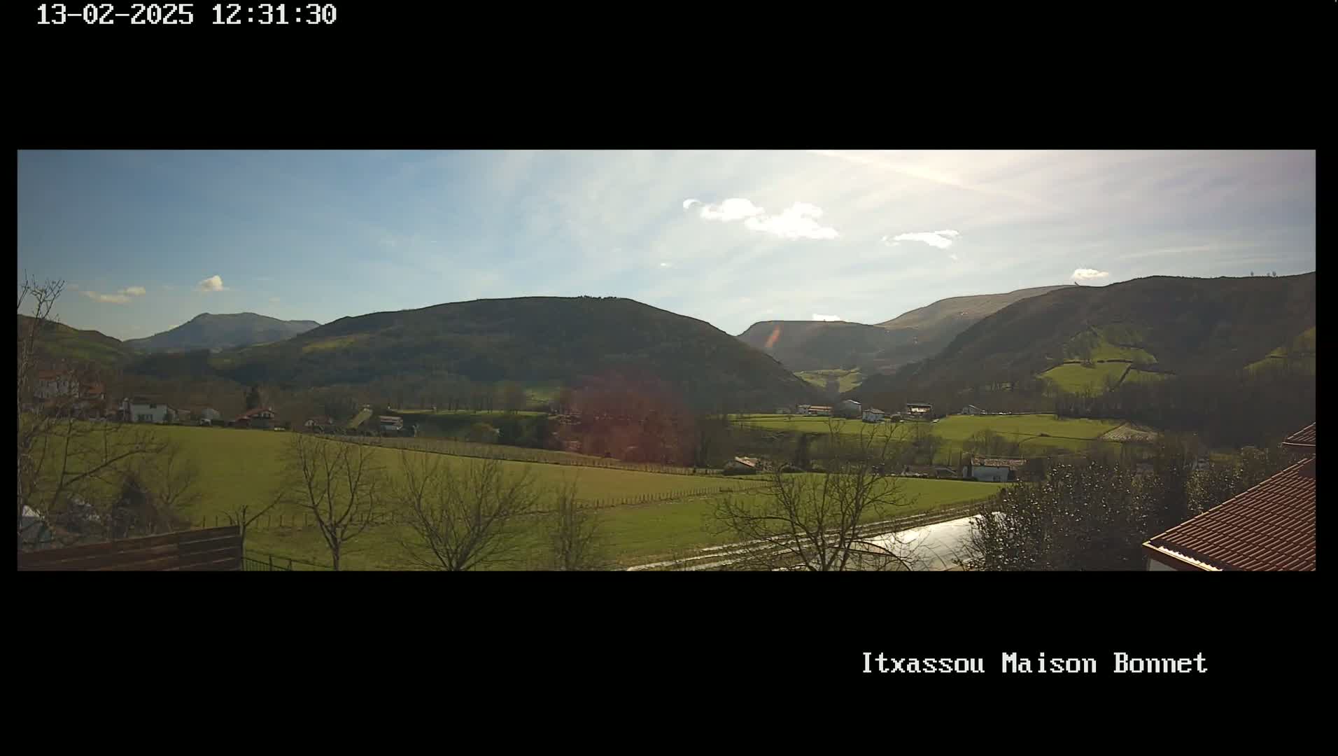 A sunny day reveals a verdant valley dotted with houses and trees, nestled between rolling hills under a mostly clear sky.