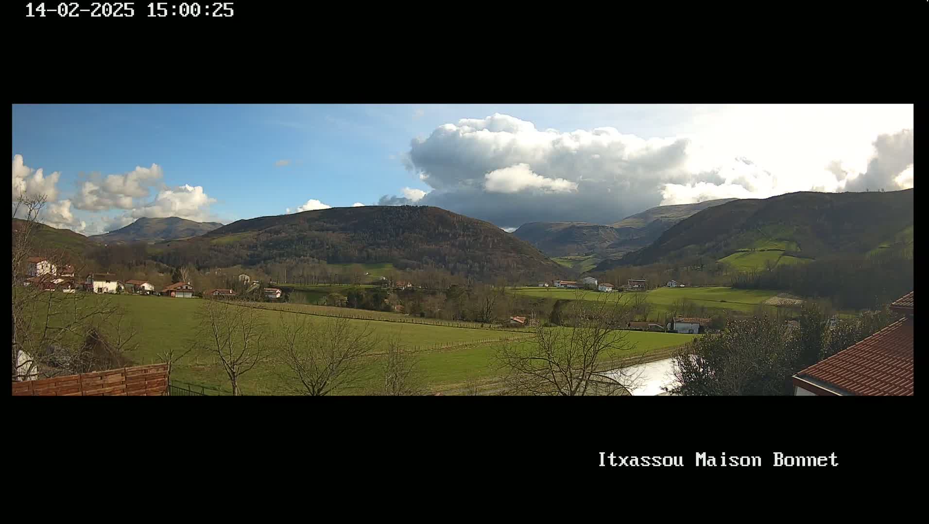 A partly cloudy day reveals a valley with green fields, a few houses, and bare winter trees, nestled among rolling hills.