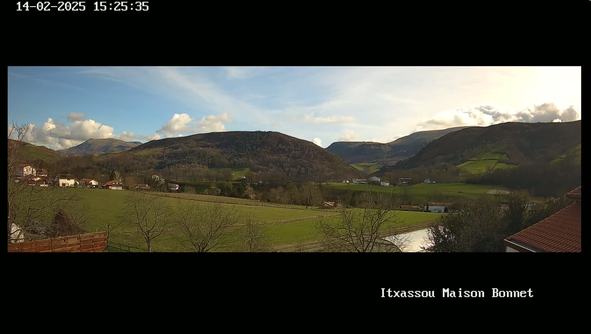 A partly cloudy day reveals a sprawling green valley dotted with houses and trees, backed by rolling hills and mountains.