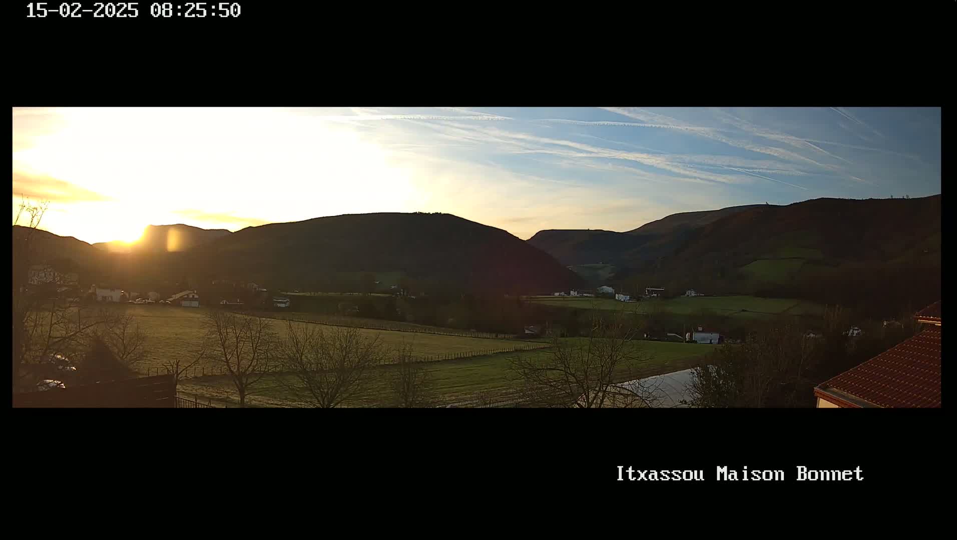 A sunrise illuminates a valley with fields and sparsely scattered houses, nestled between dark, rolling hills under a mostly clear sky.