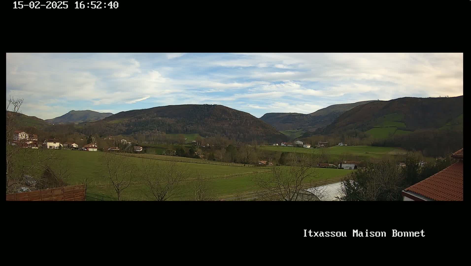 A partly cloudy day reveals a tranquil valley scene with rolling green hills, sparsely scattered houses, and leafless trees.
