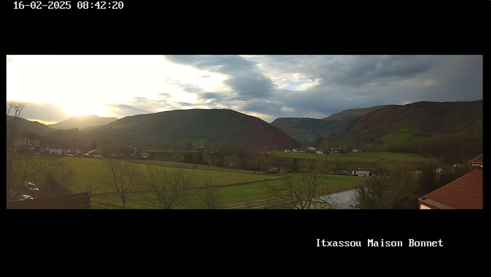 A sunlit valley with green fields and scattered houses nestled amongst rolling hills under a partly cloudy sky.