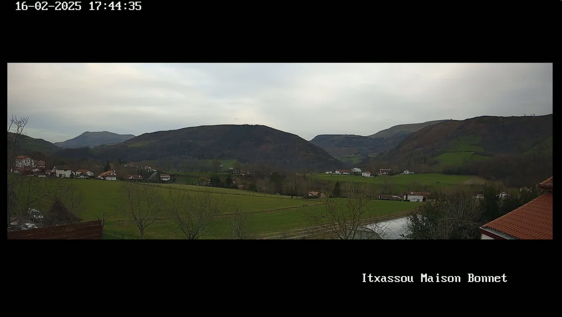 A panoramic view of a rural valley under an overcast sky shows a collection of houses nestled among green fields and rolling hills.