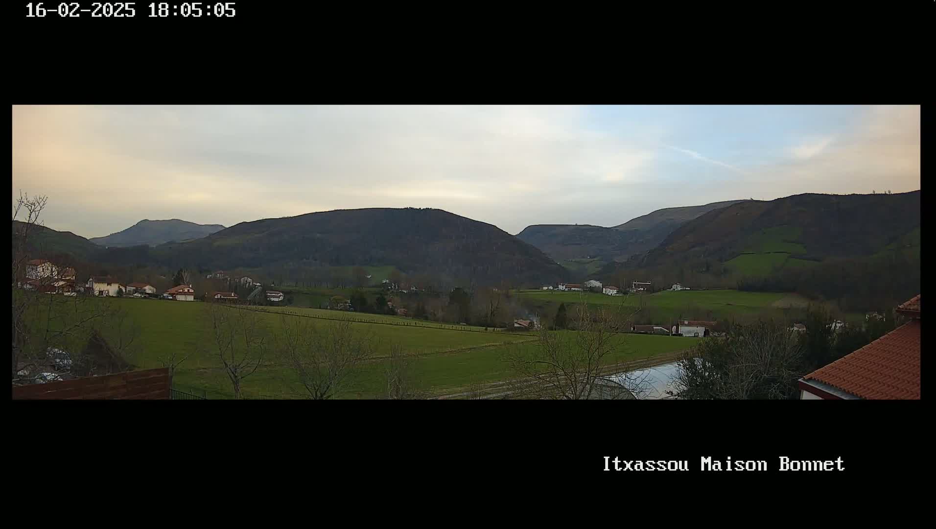 A pastoral valley scene under a cloudy, twilight sky shows a small village nestled among rolling green hills and a few trees.