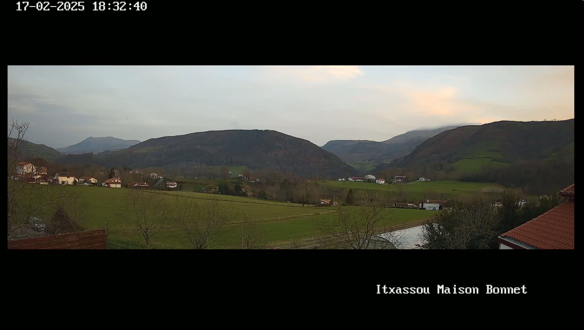 A panoramic view of a valley with scattered houses and farmland under a cloudy sky, nestled amongst rolling hills and mountains.