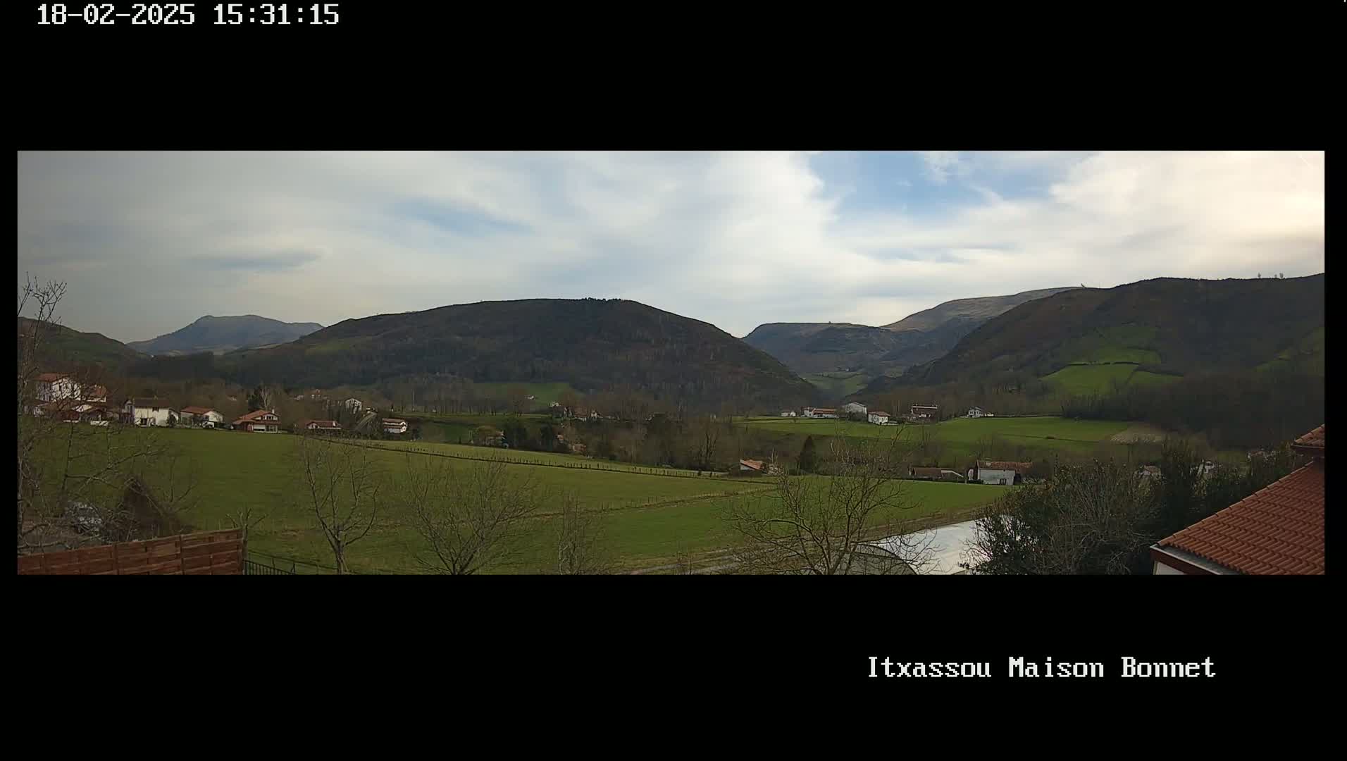A mostly cloudy day reveals a sprawling green valley dotted with houses and bordered by rolling, dark-green hills.