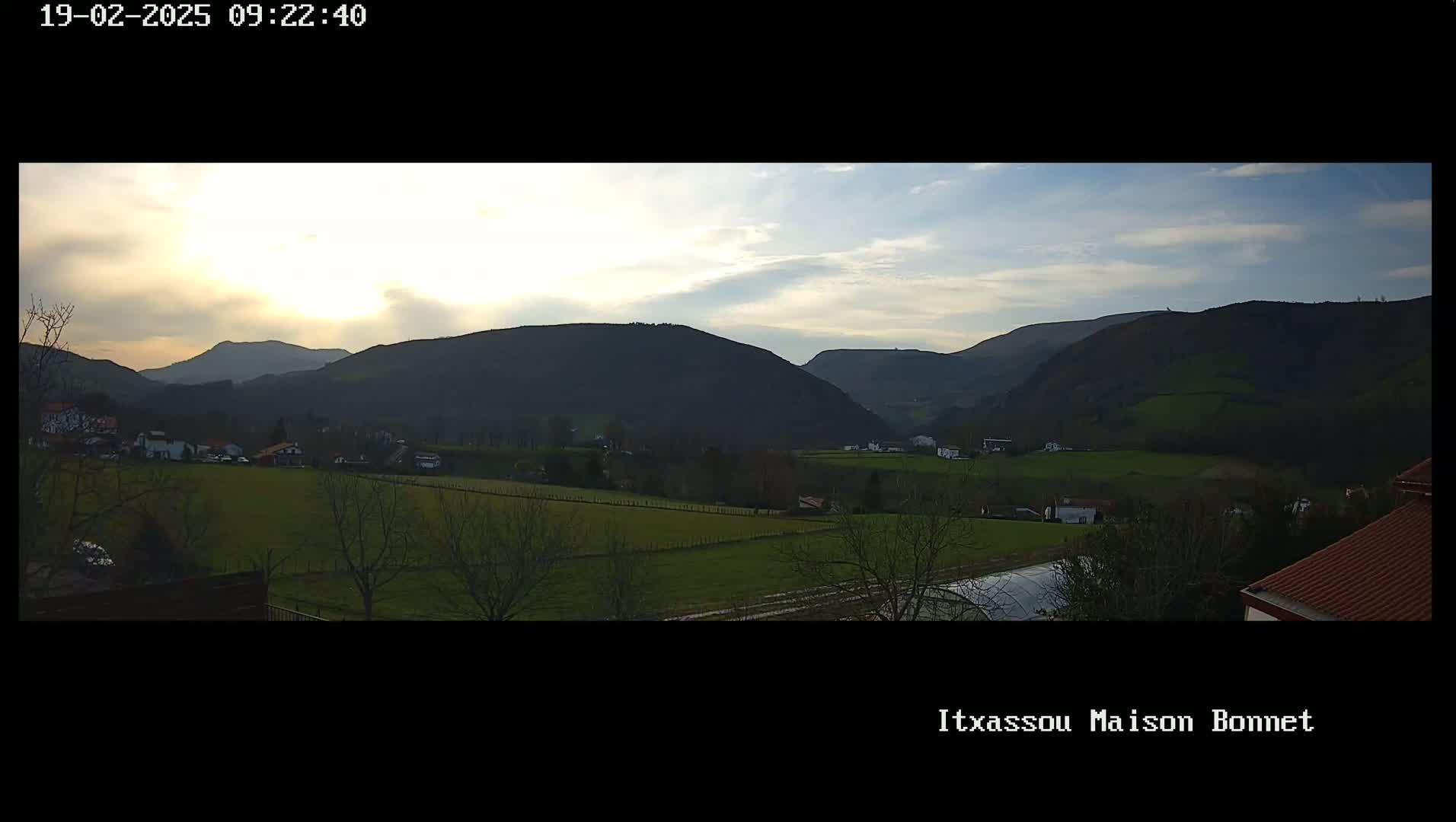 A sunlit valley dotted with houses and farmland is nestled between rolling hills under a partly cloudy sky.