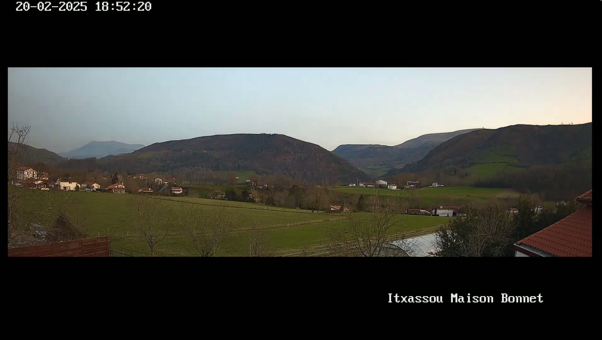 A panoramic view of a rural landscape under a clear, light-grey sky shows a valley with scattered houses and farm fields, bordered by dark-green hills and mountains in the distance.