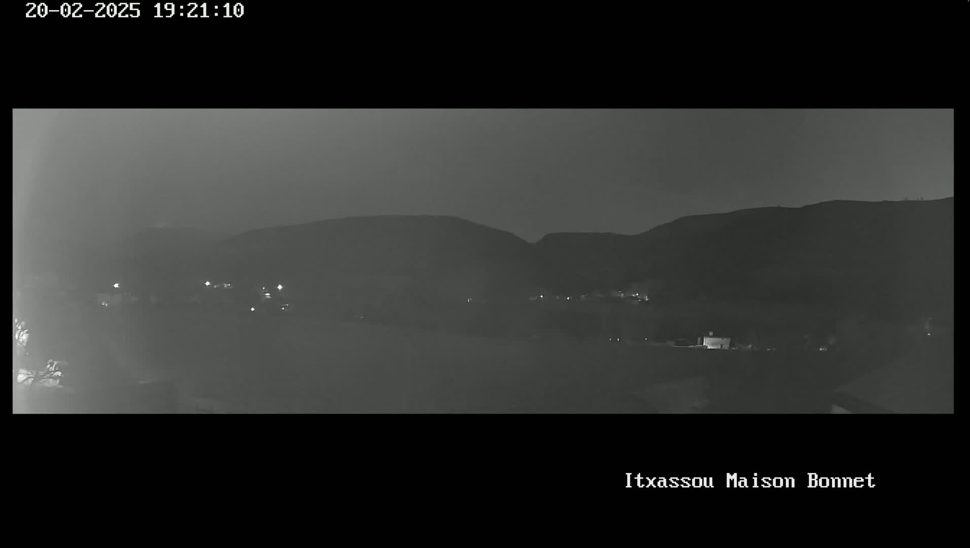 A nighttime, grayscale image shows a dark, hilly landscape with a few faint lights scattered across the lower portion.