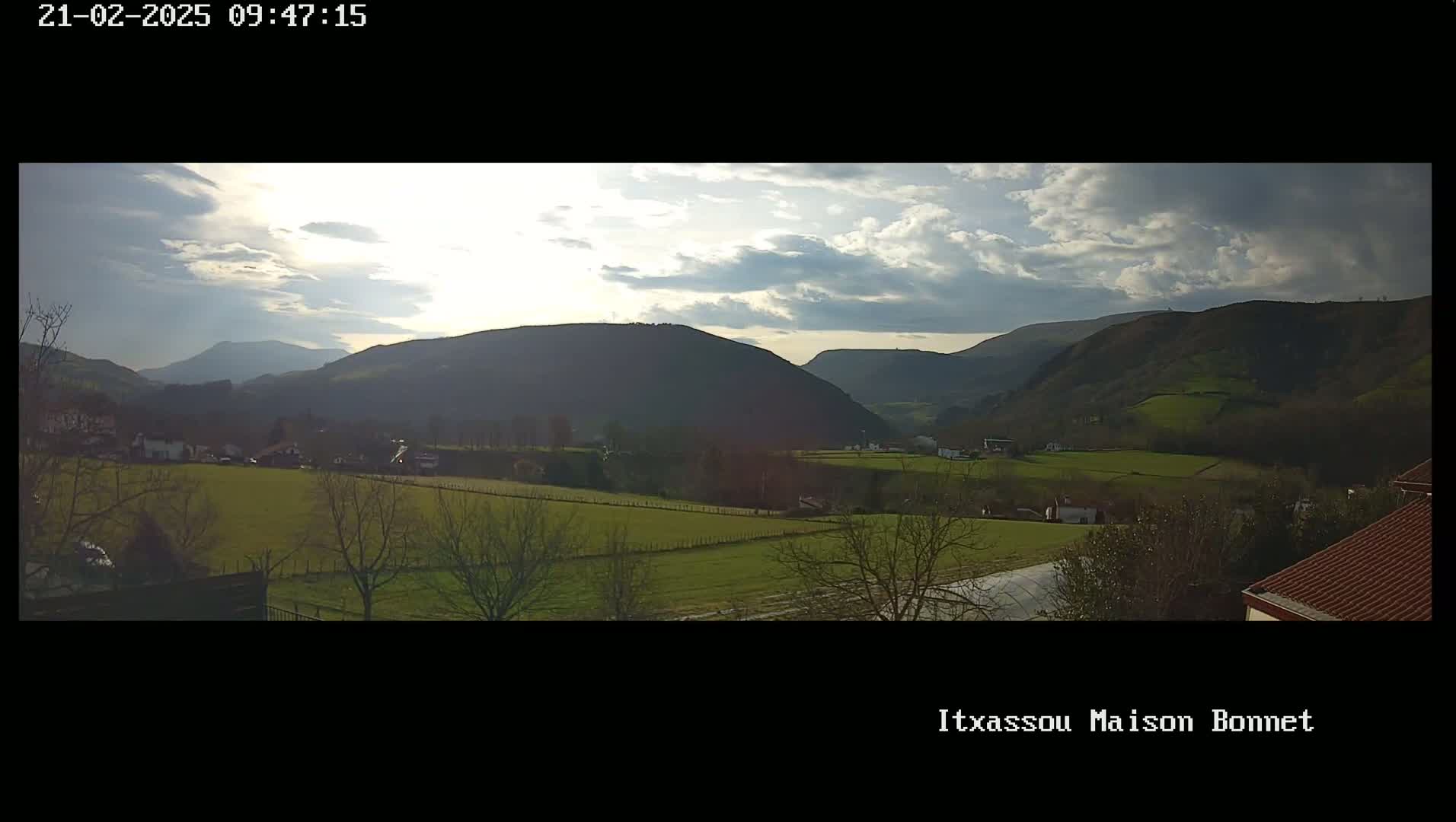 A partly cloudy day reveals a valley of green fields and low hills, with larger mountains in the background.