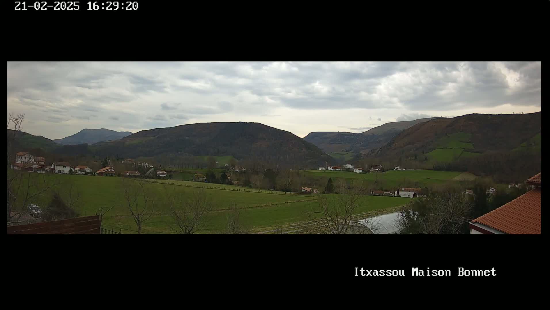 A sprawling green valley dotted with houses and farmlands is nestled between rolling hills under a cloudy sky.