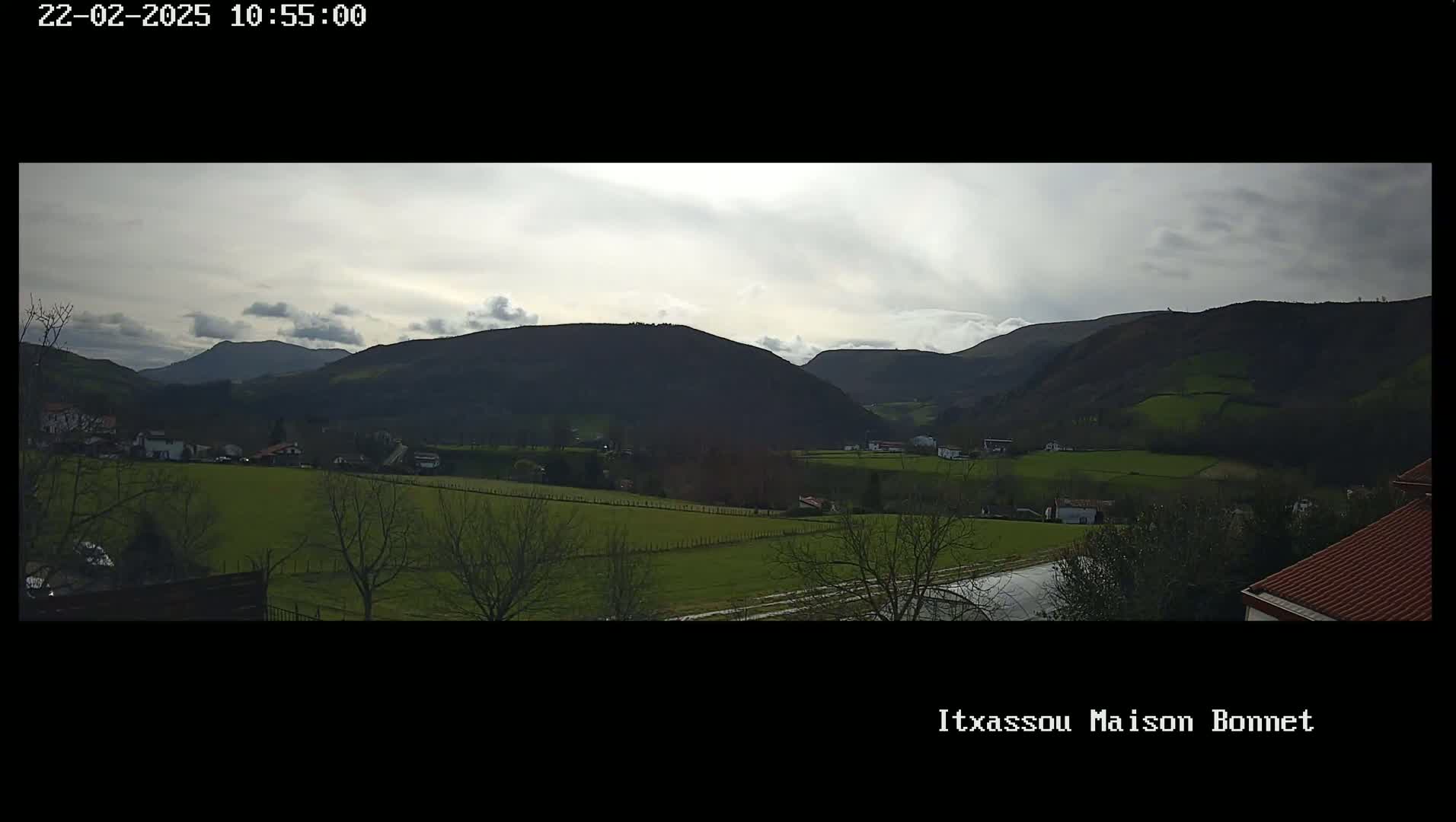 A panoramic view of a valley with rolling green hills and a few buildings under a cloudy sky.