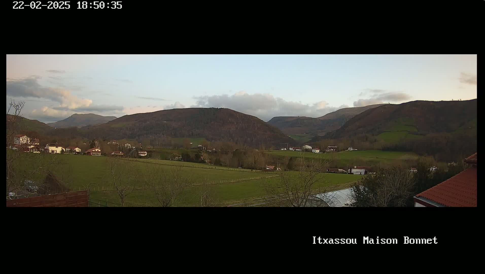 A panoramic view of a rural valley under a partly cloudy sky shows several houses nestled amongst green fields and dark-green hills.