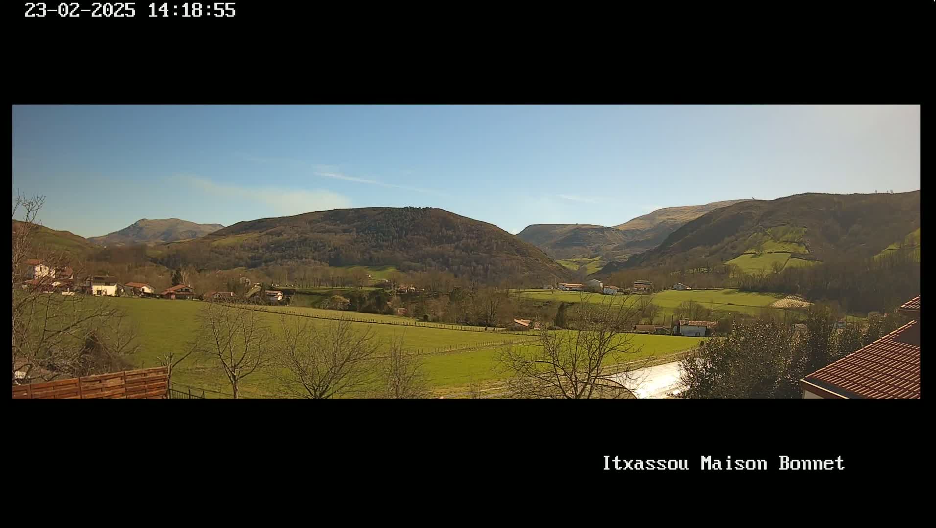 A sunny view of a valley with green fields, scattered houses, and bare trees, nestled between rolling hills under a clear blue sky.