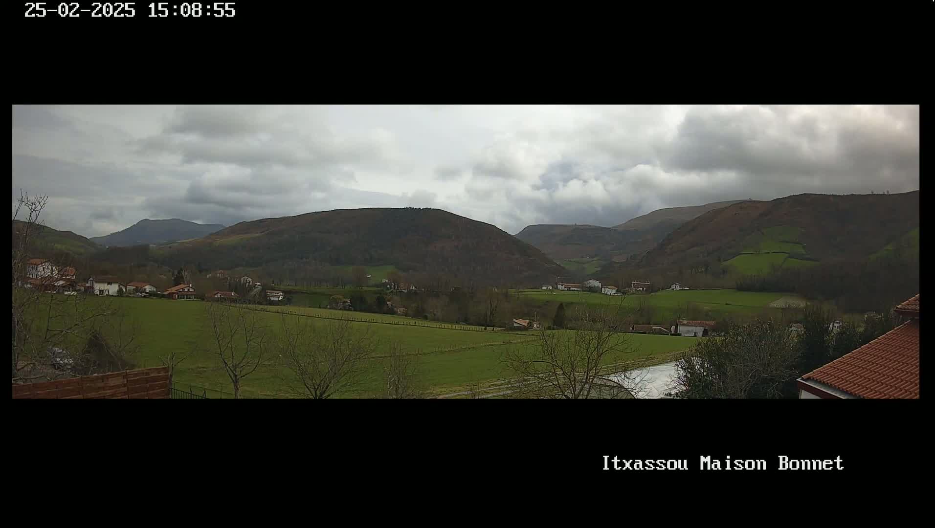 A verdant valley dotted with houses stretches beneath a cloudy sky, rolling hills rising in the background.