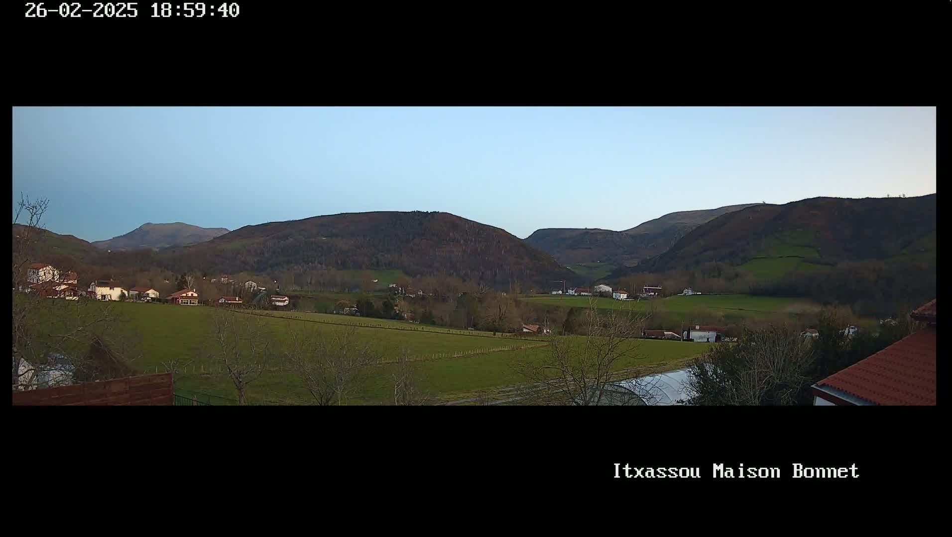 A panoramic view of a valley with a small village nestled amongst green fields and brown hills under a clear, light-blue sky.