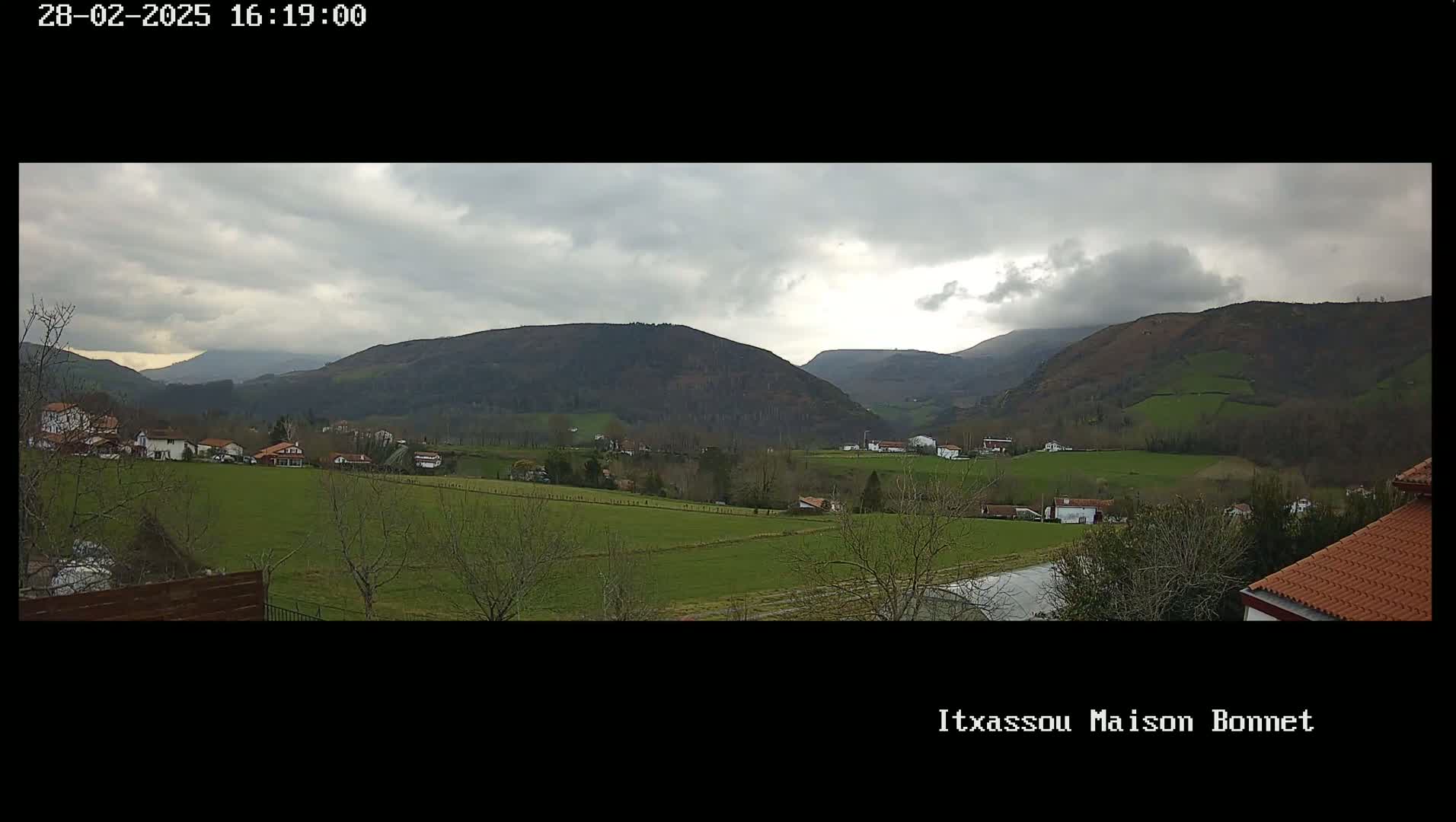 A sprawling green valley dotted with houses and trees under a cloudy sky, backed by rolling hills.