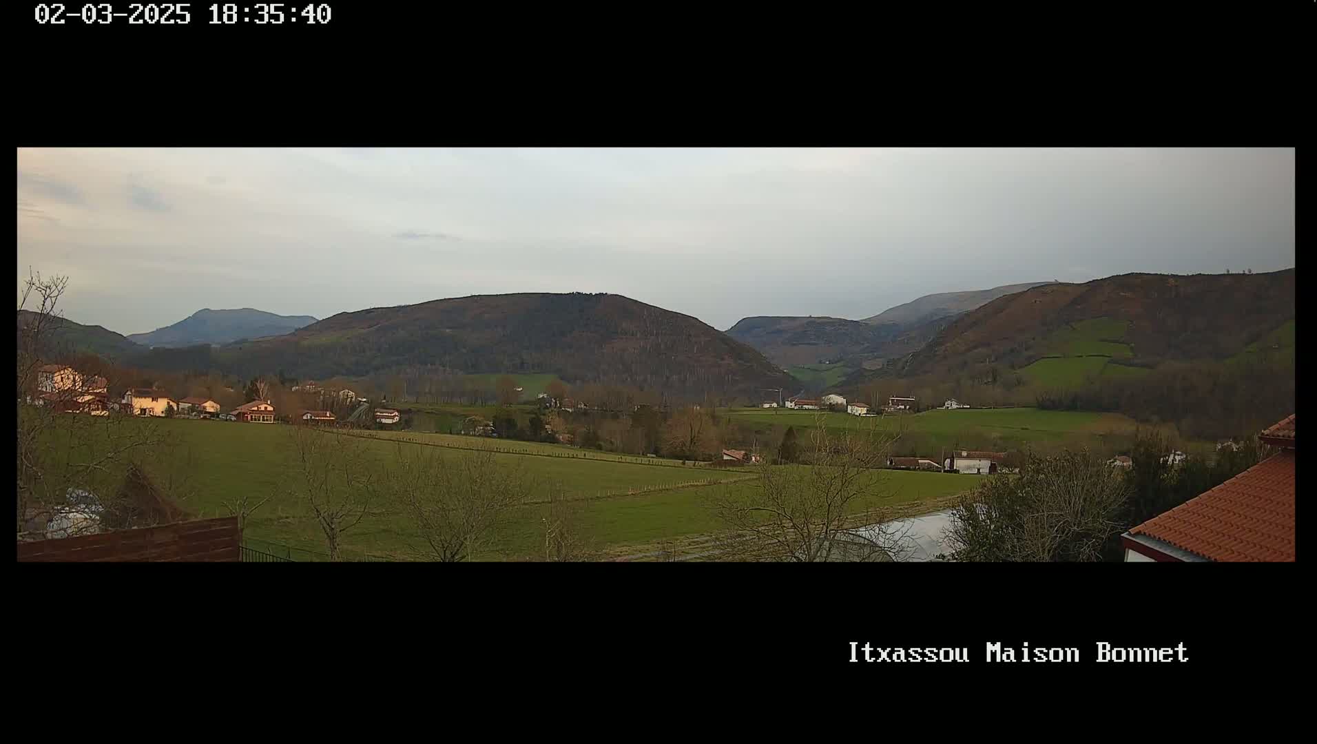 A panoramic view of a rural valley under an overcast sky shows a scattering of houses and fields nestled among rolling hills.