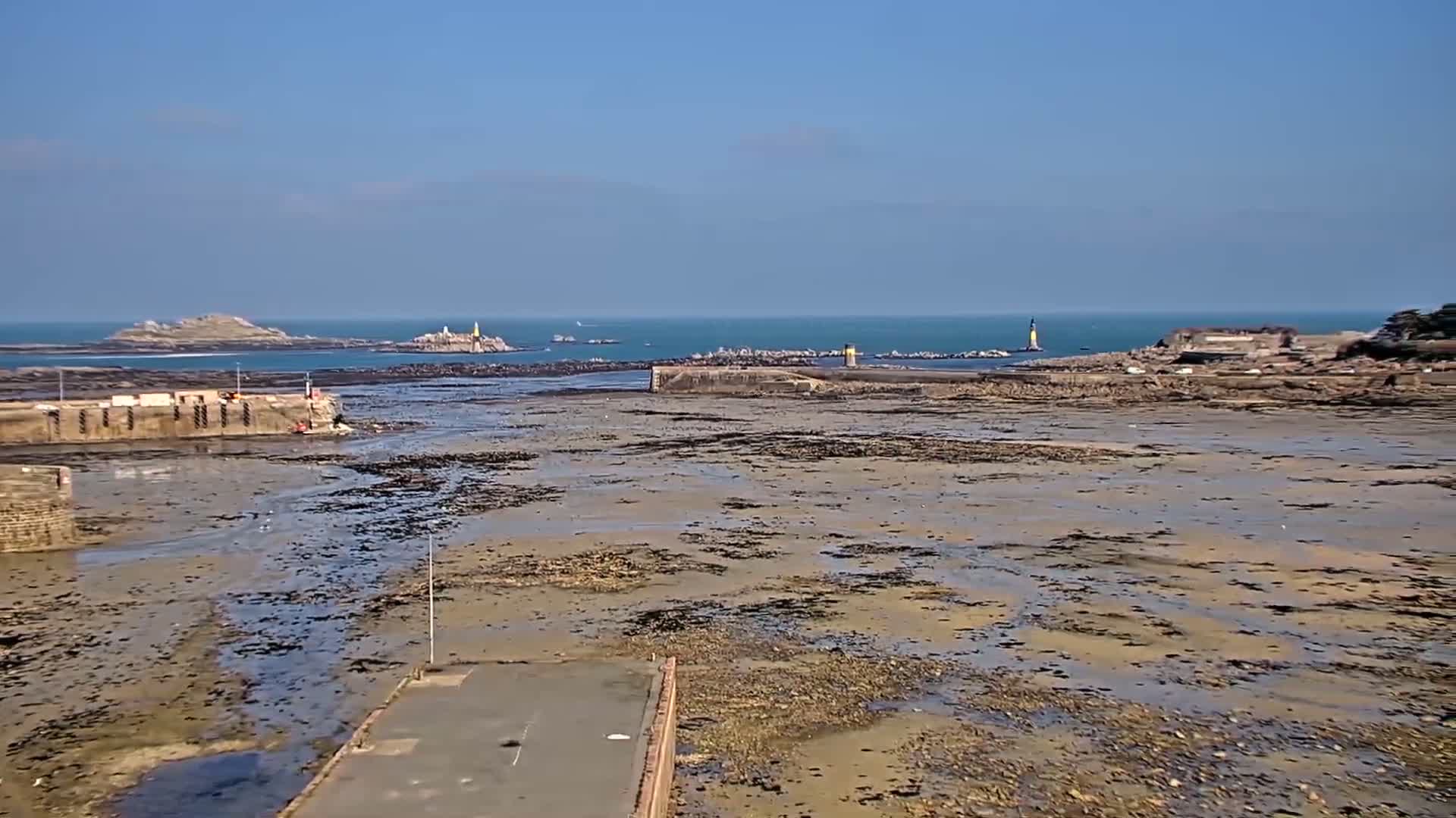 A harbor with exposed mudflats at low tide, under a clear blue sky, shows distant islands and lighthouses.