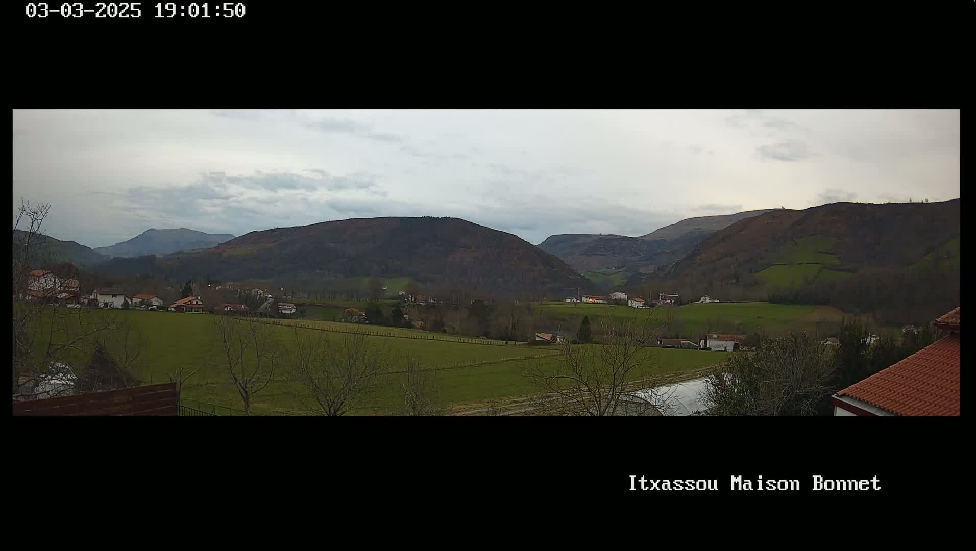 A rural valley, under a cloudy sky, shows a scattering of houses amongst green fields and dark, rolling hills.