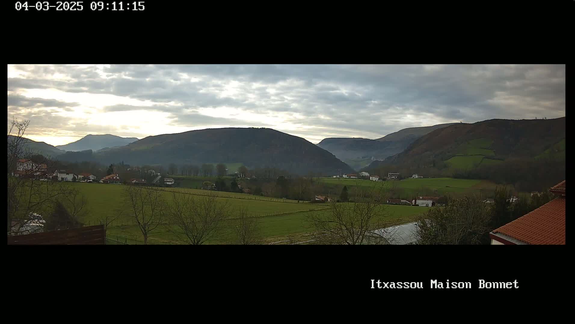 A partly cloudy sky overlooks a valley of green fields and small houses nestled amongst bare winter trees and rolling hills.