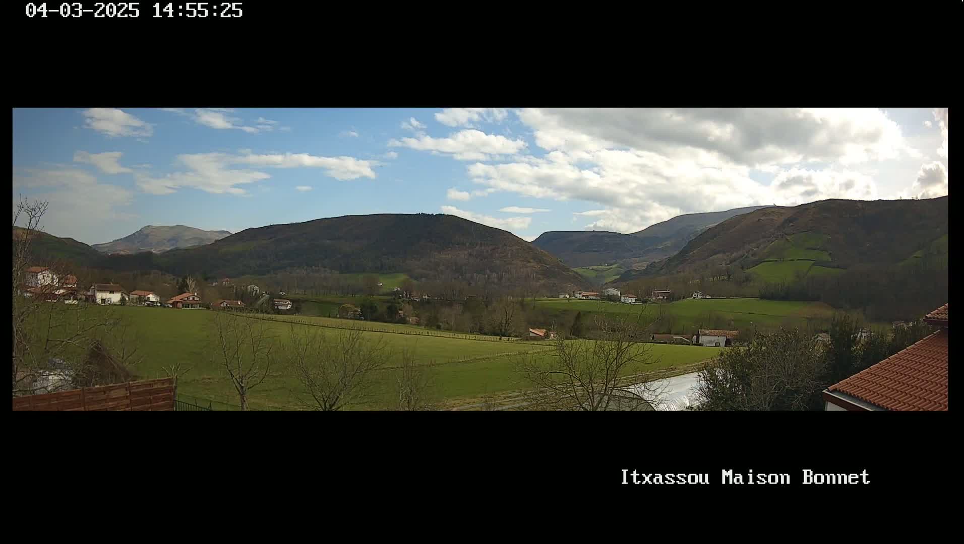 A partly cloudy day reveals a valley of green fields and small houses nestled among low, tree-covered hills and mountains.