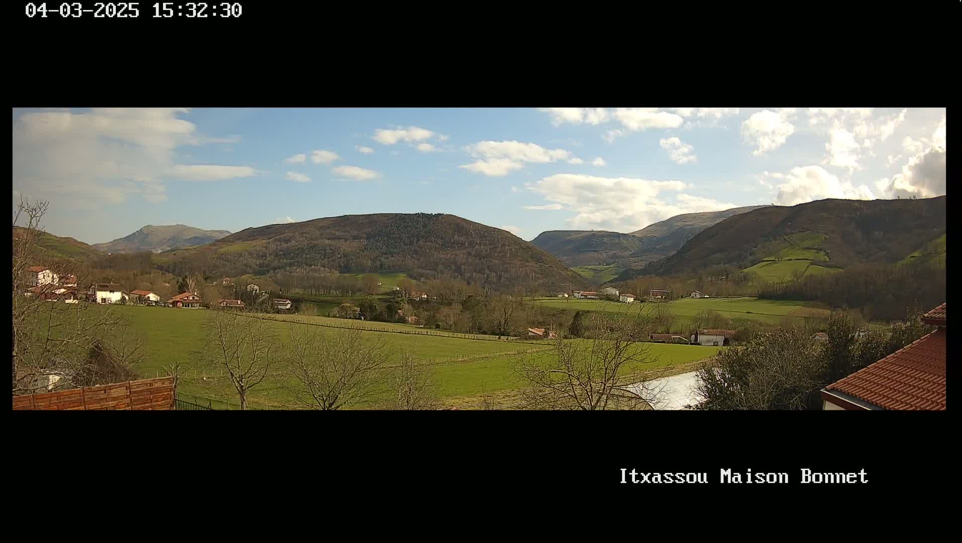 A partly cloudy day reveals a valley of green fields and sparse trees nestled between rolling, wooded hills.