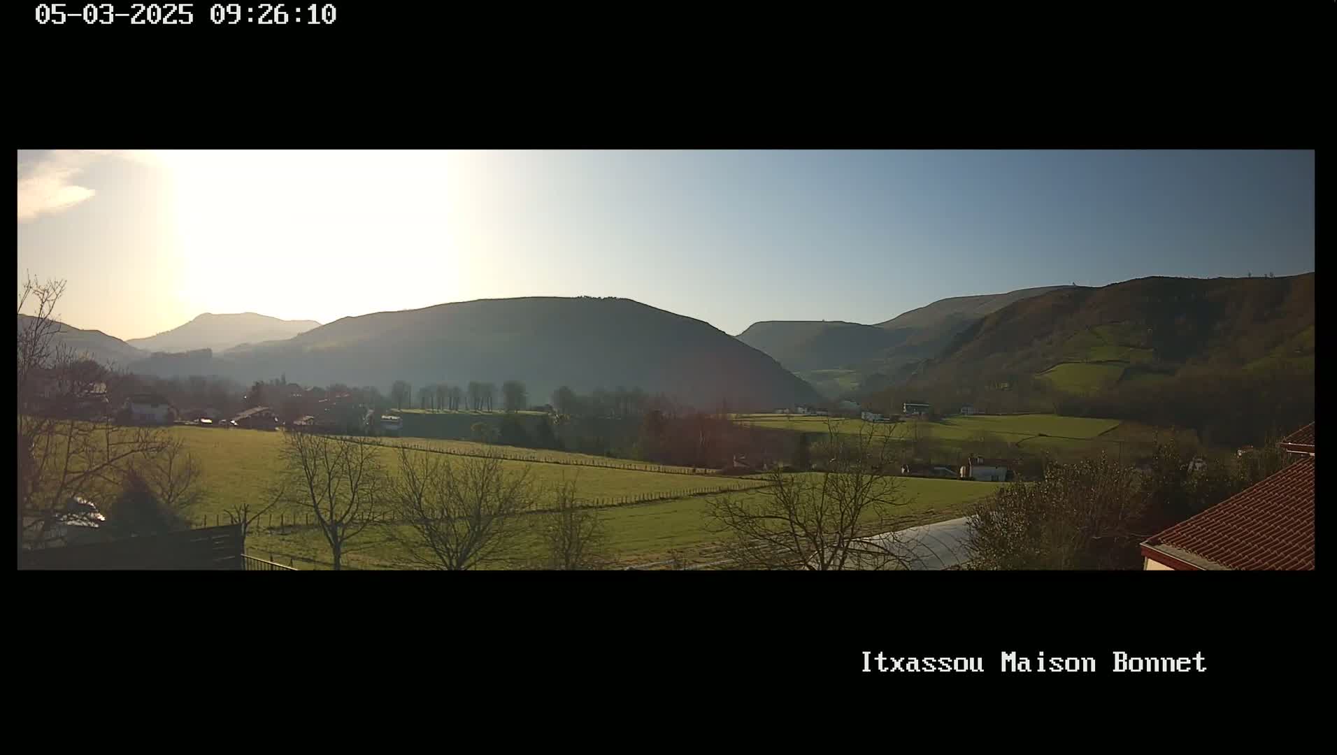 A panoramic view of a sunlit valley dotted with farmlands and villages, nestled among rolling hills under a clear sky.