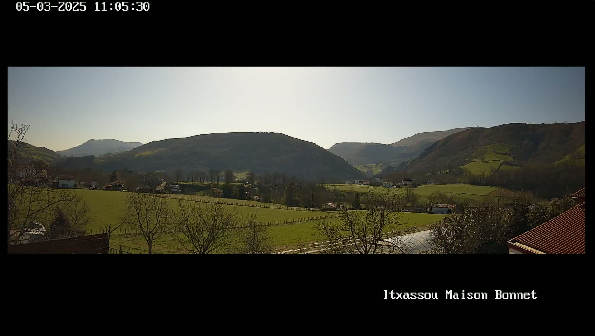 A sunny view of a valley with green fields, bare trees, and distant rolling hills.