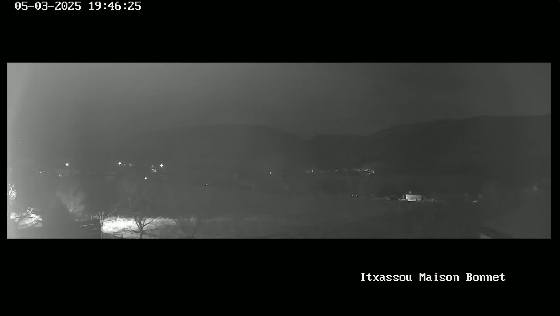 A nighttime, low-light view shows a dimly lit landscape with hills in the background and sparse, indistinct structures and trees in the foreground.