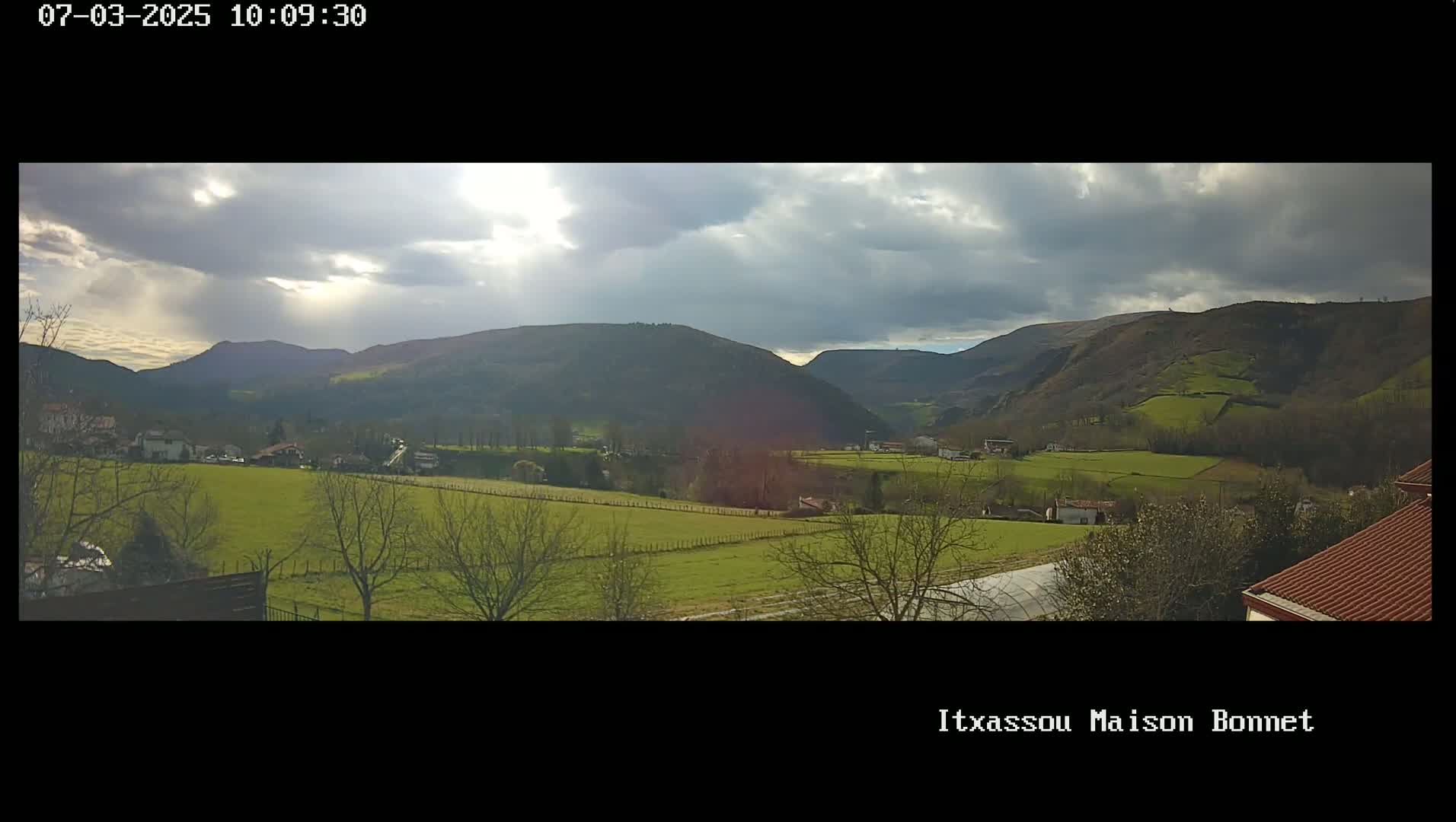 A partly cloudy day reveals a valley of green fields and sparse trees, nestled between rolling hills.