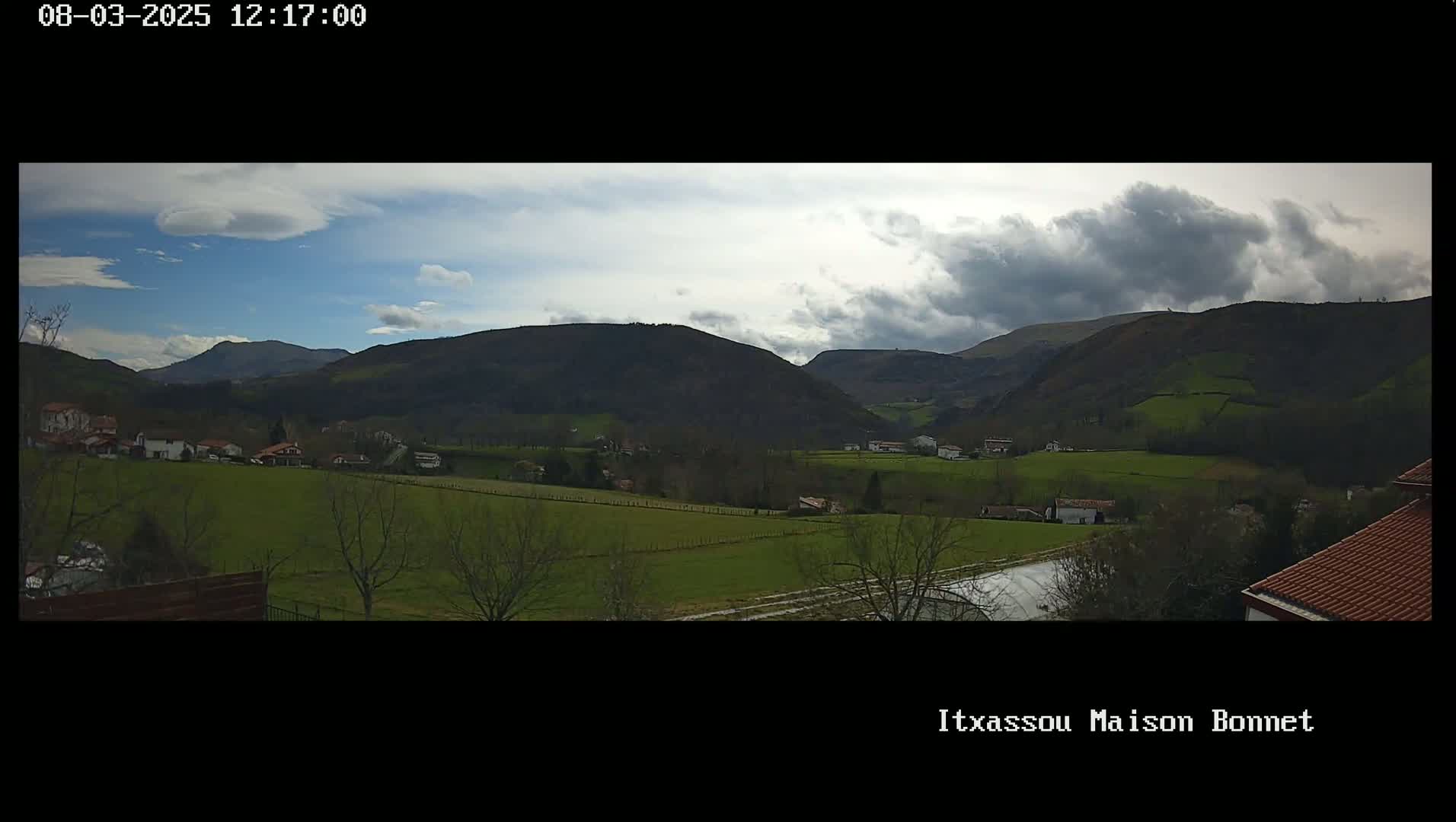 A partly cloudy day reveals a valley of green fields and scattered houses nestled between dark, rolling hills.