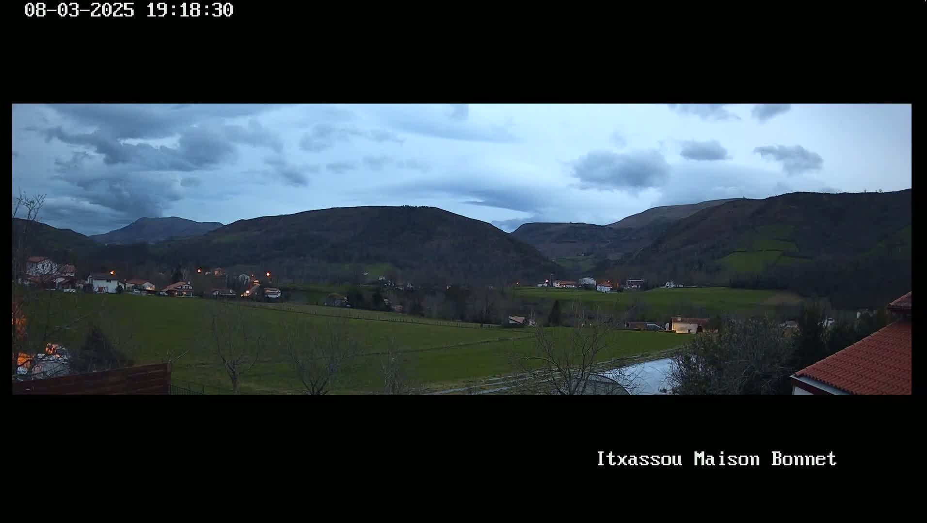 A rural village nestled in a valley between dark hills under a cloudy twilight sky.