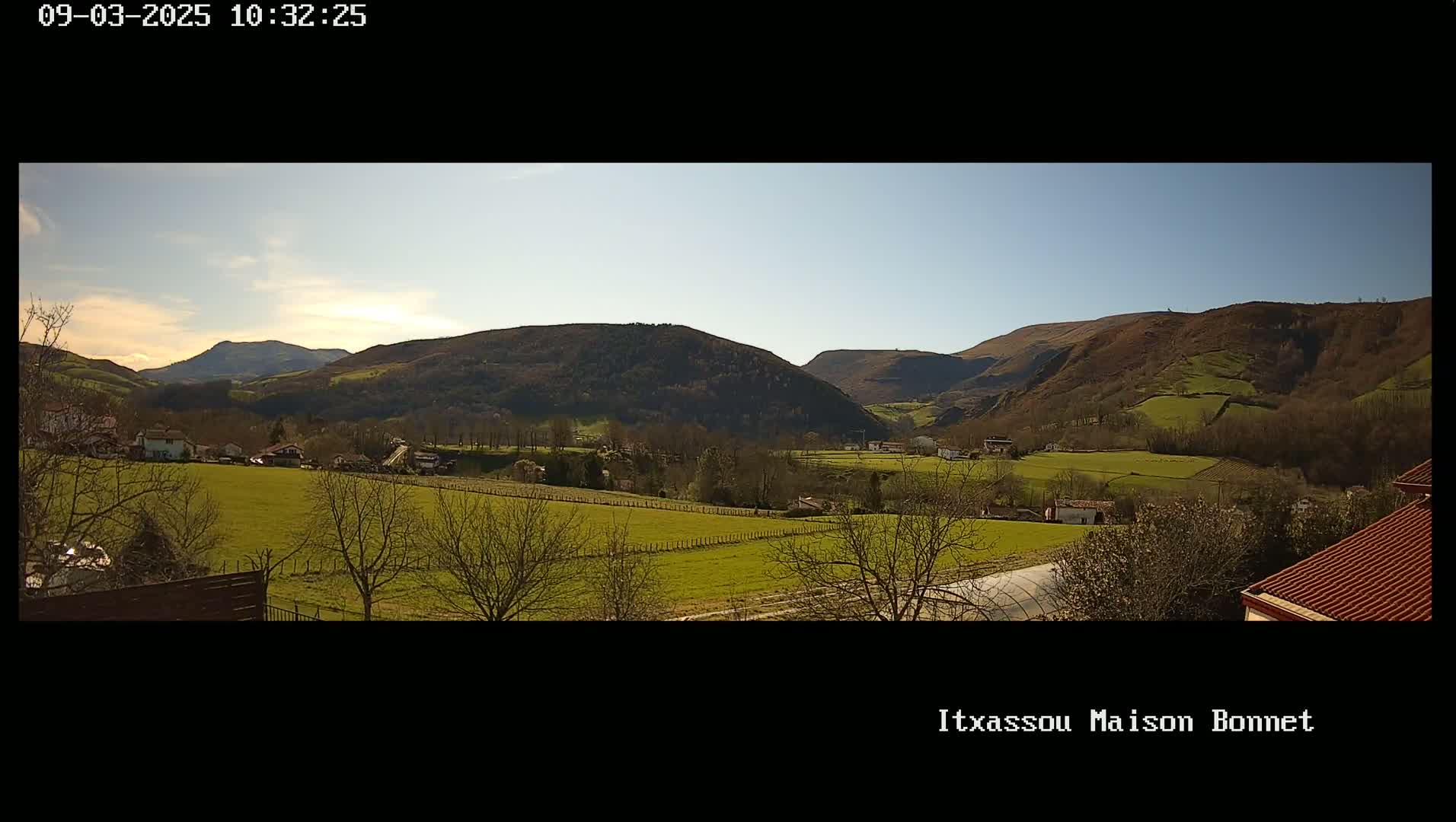 A sunny day reveals a valley of green fields and sparse trees nestled between rolling, brown-green hills.