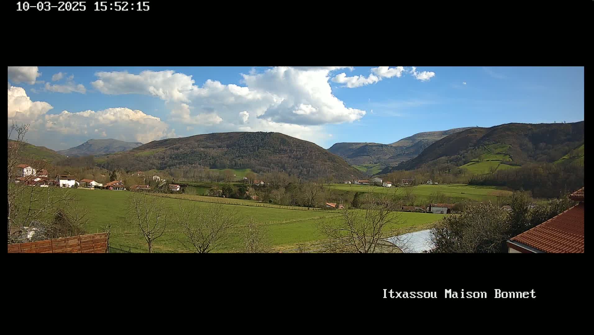 A partly cloudy, sunny day reveals a sprawling green valley dotted with houses and trees, nestled between rolling hills and mountains.