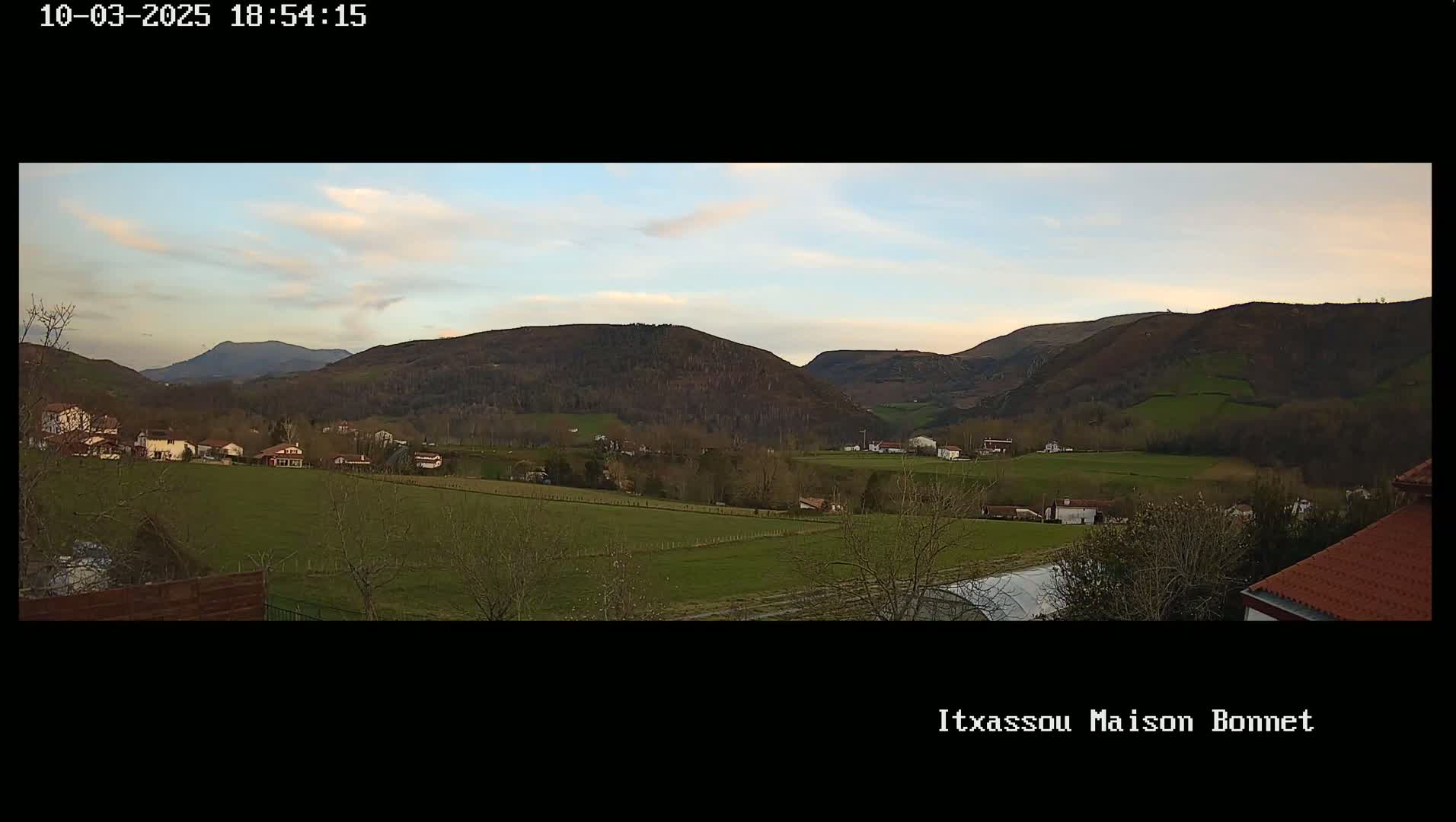 A tranquil valley scene under a partly cloudy sky shows a scattering of houses and farmlands nestled amongst rolling hills.