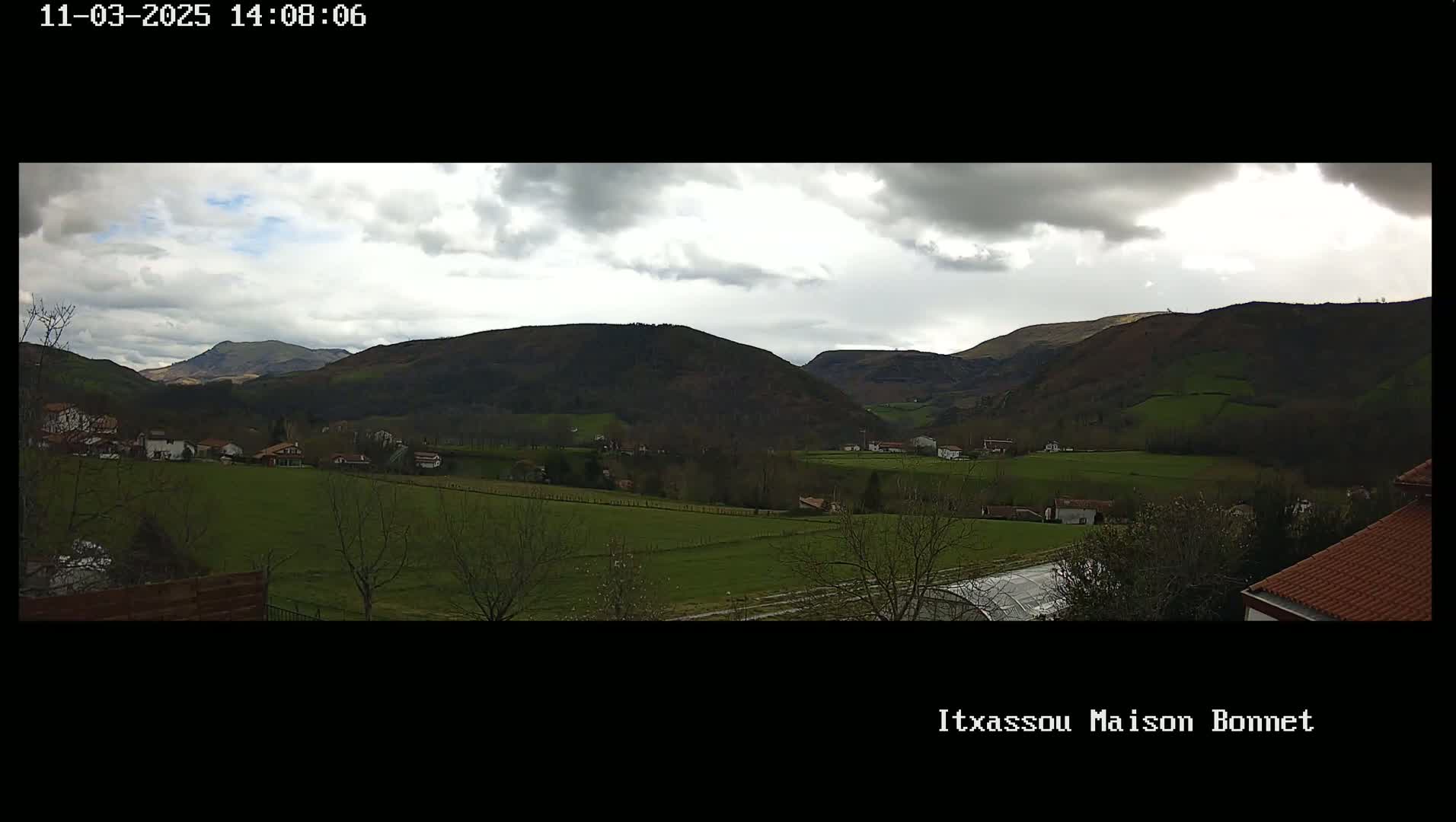 A sprawling green valley dotted with houses and trees under a partly cloudy sky is nestled between rolling hills.