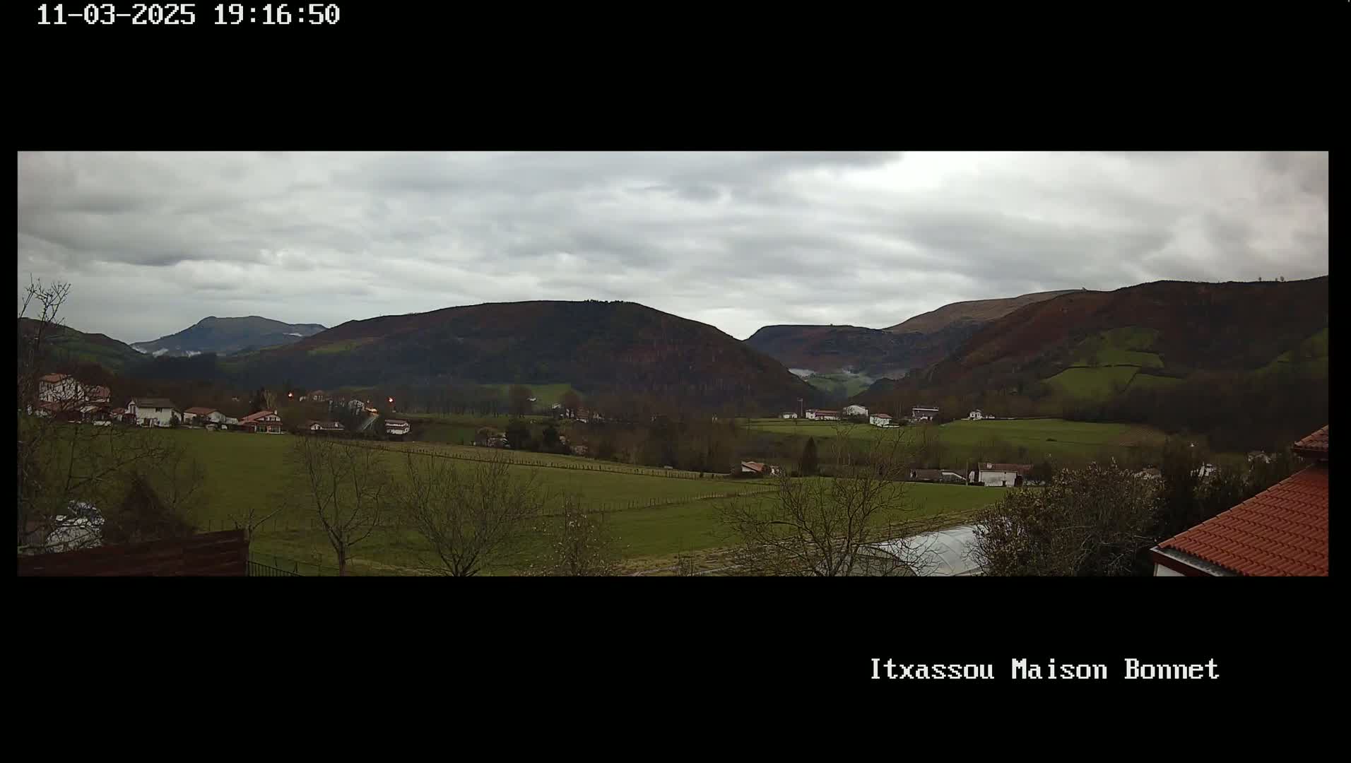 A sprawling green valley dotted with houses sits beneath a cloudy sky, backed by dark, rolling hills.