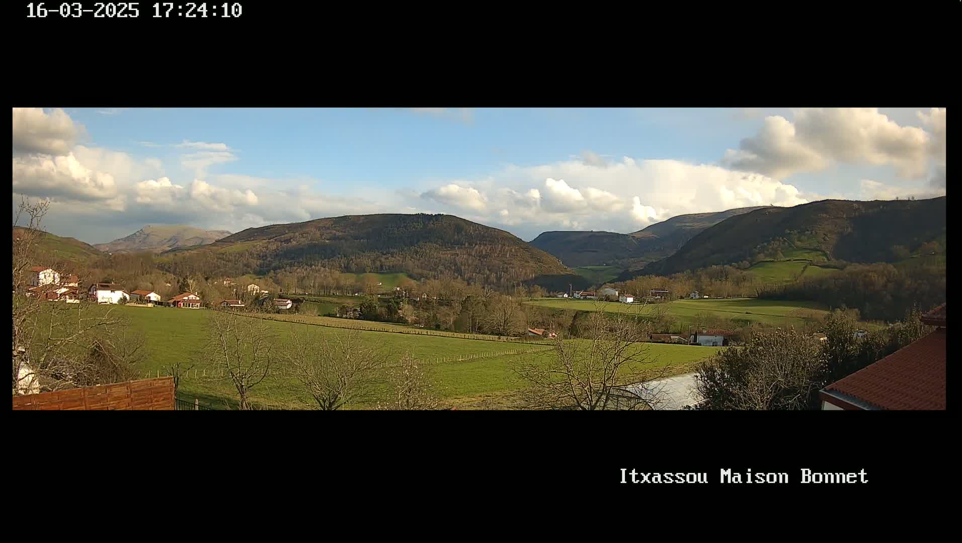 A partly cloudy daytime view of a verdant valley with a scattering of houses and trees, nestled between rolling hills.