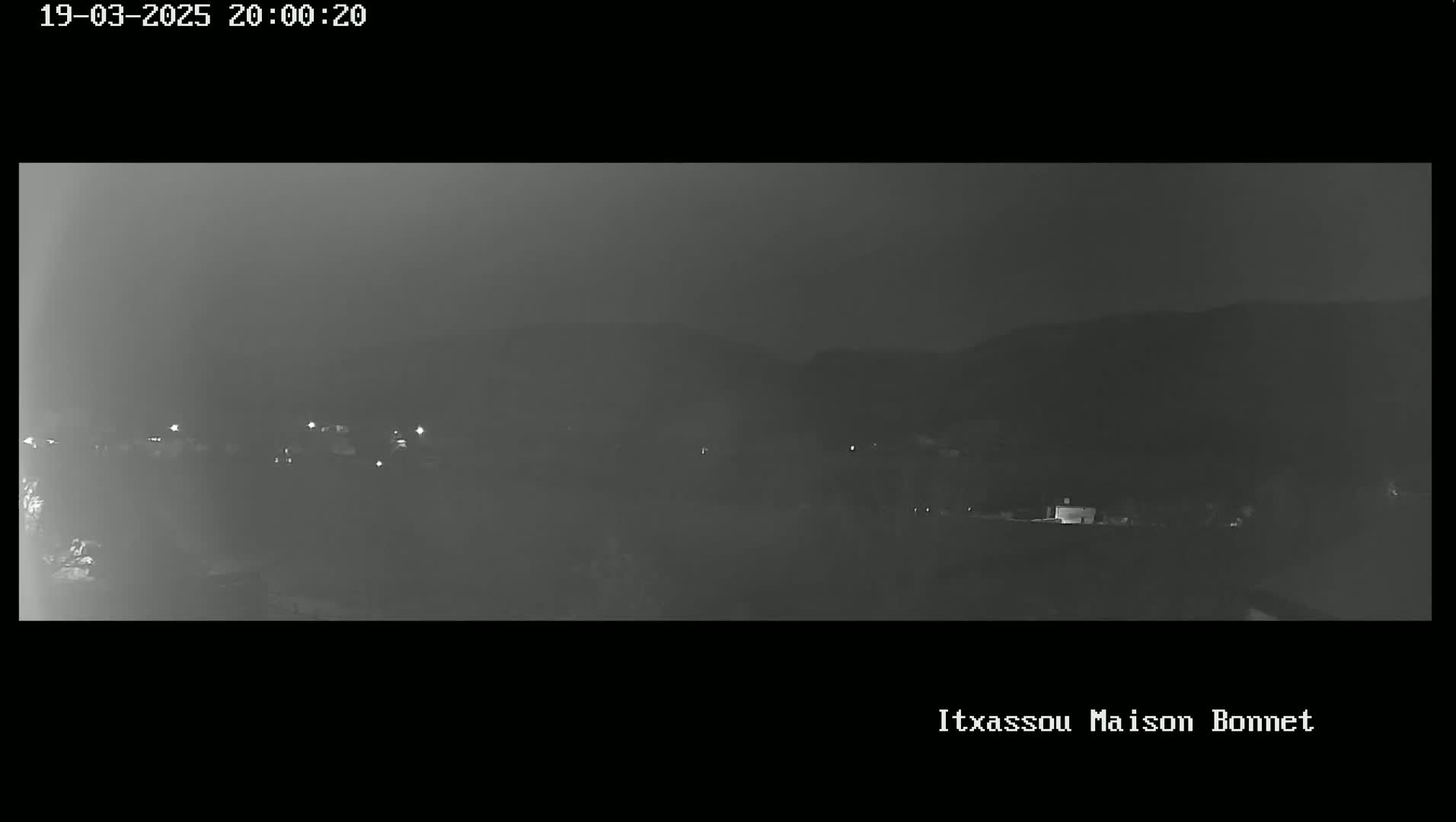 A nighttime, hazy long shot of a dimly lit town nestled in a valley, with faint lights visible against a dark hillside.