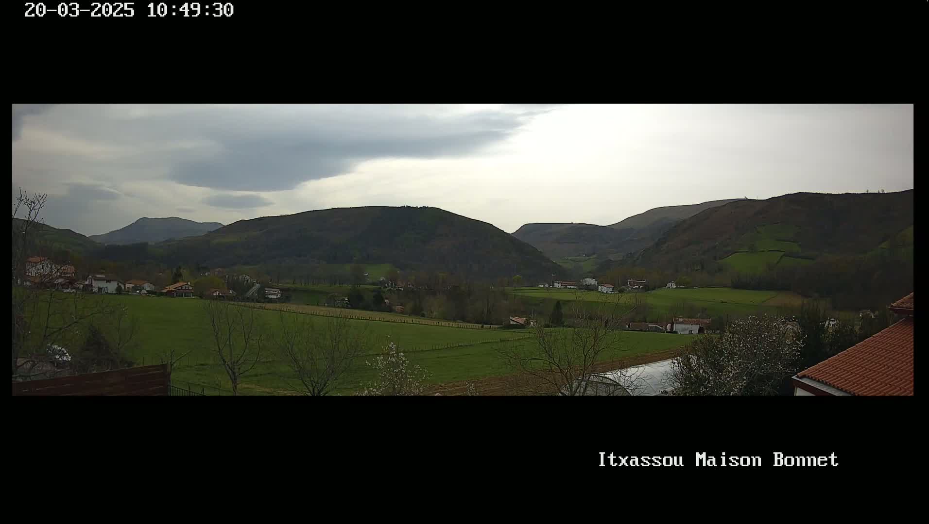 A partly cloudy day reveals a valley of green fields and small buildings nestled between rolling hills.