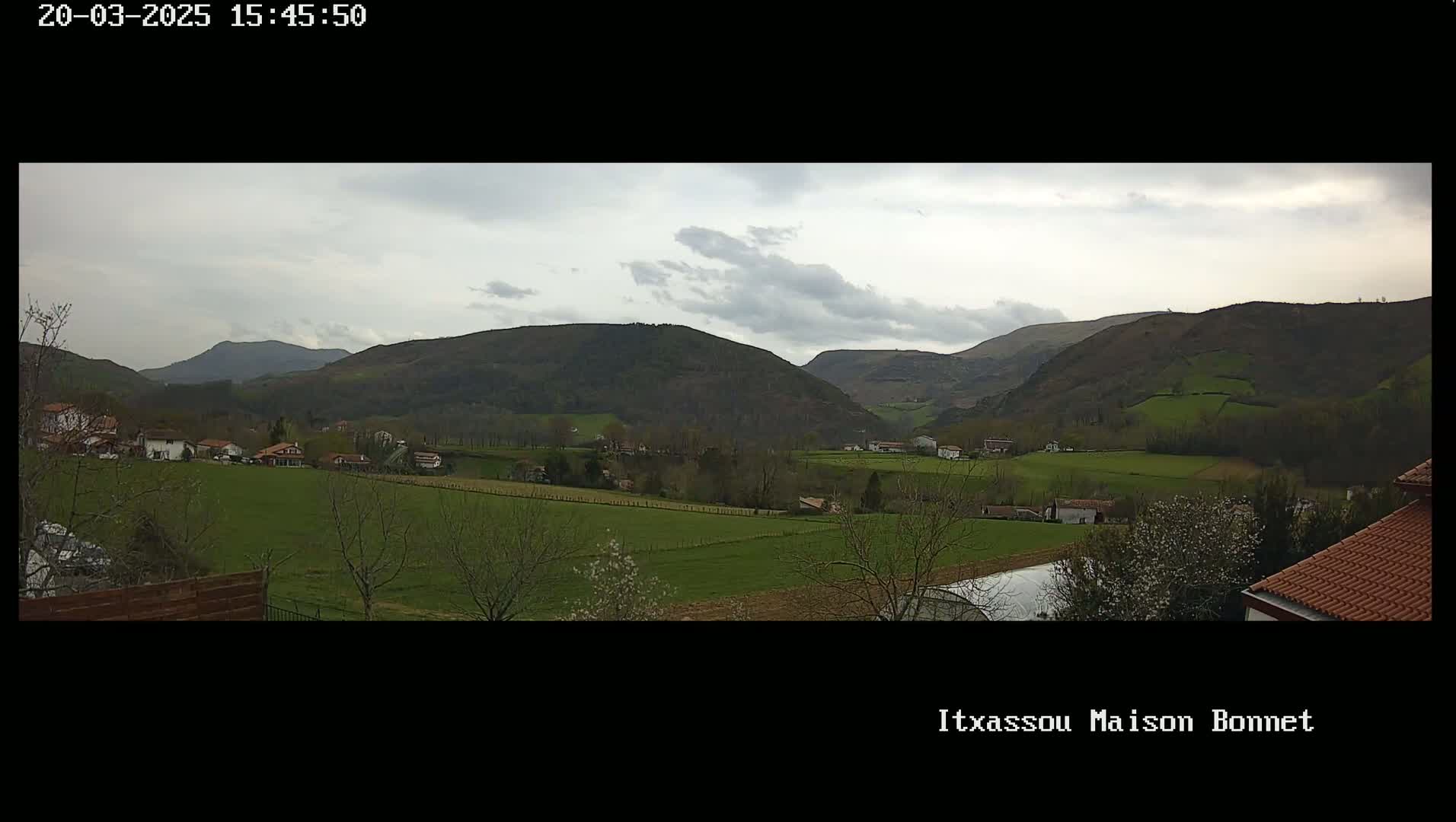 A sprawling green valley dotted with houses and farmlands, nestled beneath a cloudy sky and surrounded by rolling hills.
